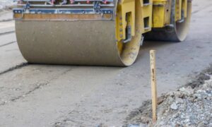 A road roller compacts a paved surface beside a wooden survey stake, showing how a construction survey helps guide grading and site layout
